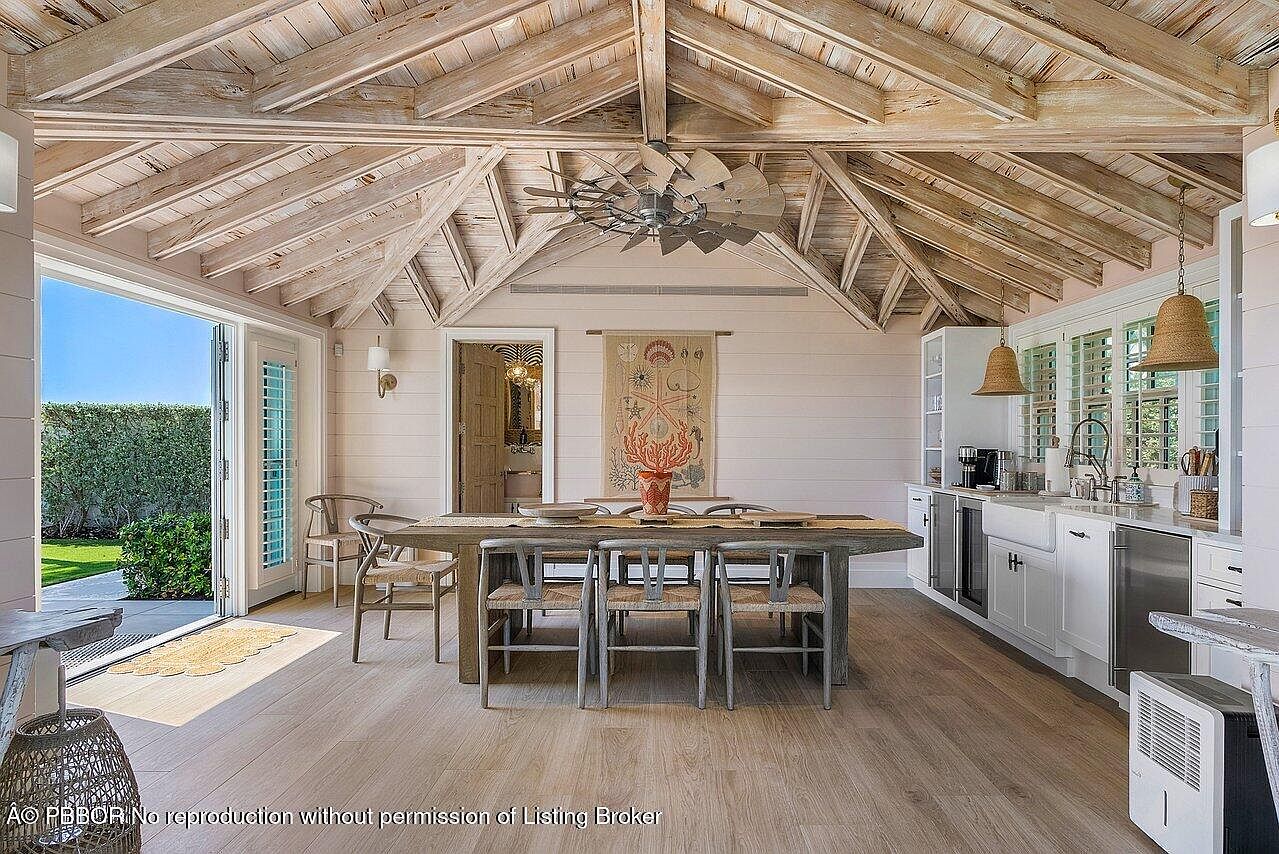 This is an interior shot of a dining room with a rustic, coastal aesthetic. The room features a large wooden dining table with seating for six, set beneath a vaulted ceiling with exposed beams and a unique windmill-style ceiling fan. A doorway leads to an outdoor area, and a kitchen area with white cabinetry and pendant lighting is visible in the background.