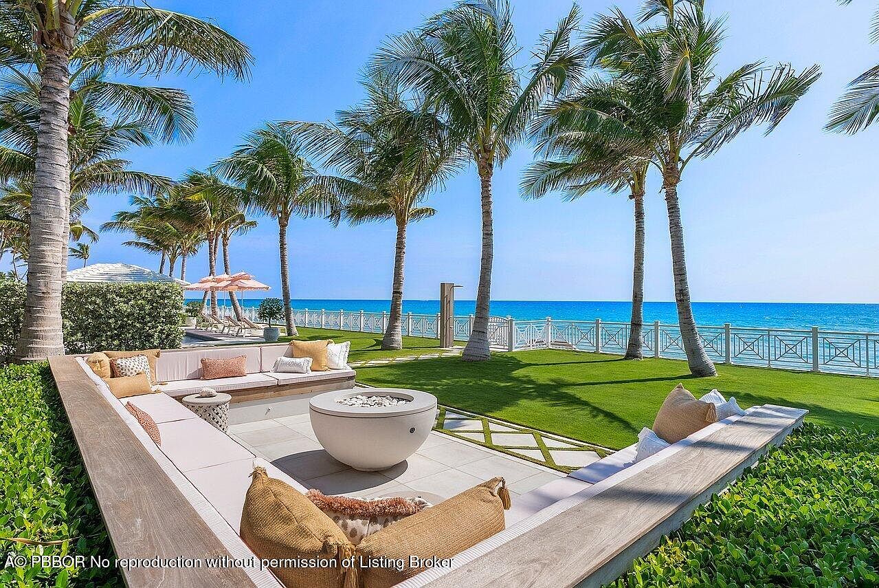 This image showcases a luxurious outdoor patio area with a stunning ocean view. The patio features a built-in seating area with plush cushions, a modern fire pit, and meticulously manicured landscaping with palm trees lining the waterfront. The overall impression is one of upscale relaxation and coastal elegance.