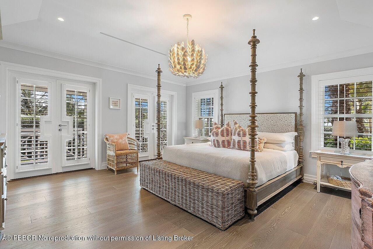 This is a bright and airy primary bedroom featuring a four-poster bed with decorative pillows and a woven bench at the foot. The room has hardwood floors, white trim, and multiple windows with plantation shutters, allowing for ample natural light. A unique chandelier hangs from the ceiling, adding a touch of elegance.