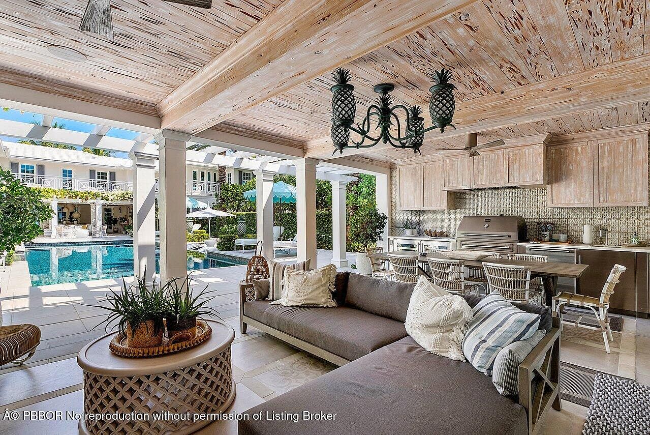 This image showcases a luxurious outdoor living space featuring a covered patio with a weathered wood ceiling and a unique pineapple chandelier. A comfortable sectional sofa is arranged around a decorative coffee table, while an outdoor kitchen and dining area are visible in the background. The patio overlooks a stunning pool and manicured gardens, creating an inviting and upscale atmosphere.