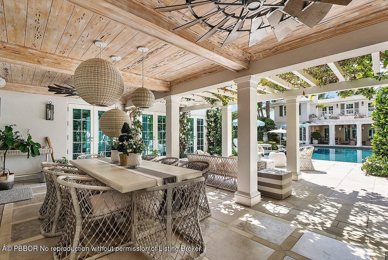 This image showcases a beautifully designed outdoor patio area, featuring a dining set with woven chairs and a large table, complemented by overhead wicker pendant lights and a rustic wooden ceiling. The space transitions seamlessly into a lounge area with comfortable seating, all overlooking a pristine pool and a well-maintained garden, creating an inviting and luxurious outdoor living experience. The view extends to a grand two-story house in the background, enhancing the property's appeal.