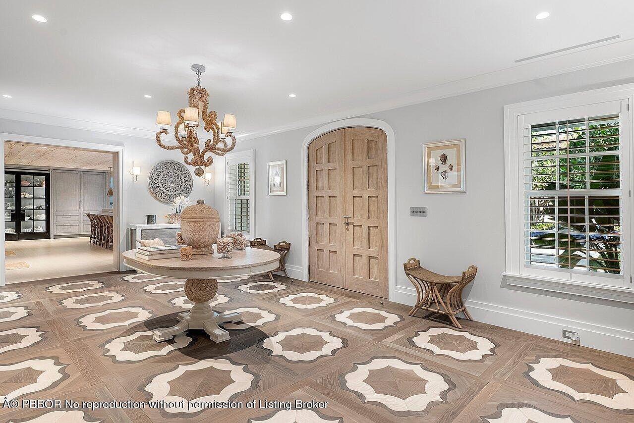 This is an interior shot of a grand entryway featuring a decorative patterned hardwood floor, a round table with decorative items, and an ornate chandelier. A wooden arched doorway and a window with shutters add architectural interest, while the neutral wall color creates a bright and inviting space. The perspective is from the center of the room, capturing the full scope of the design.