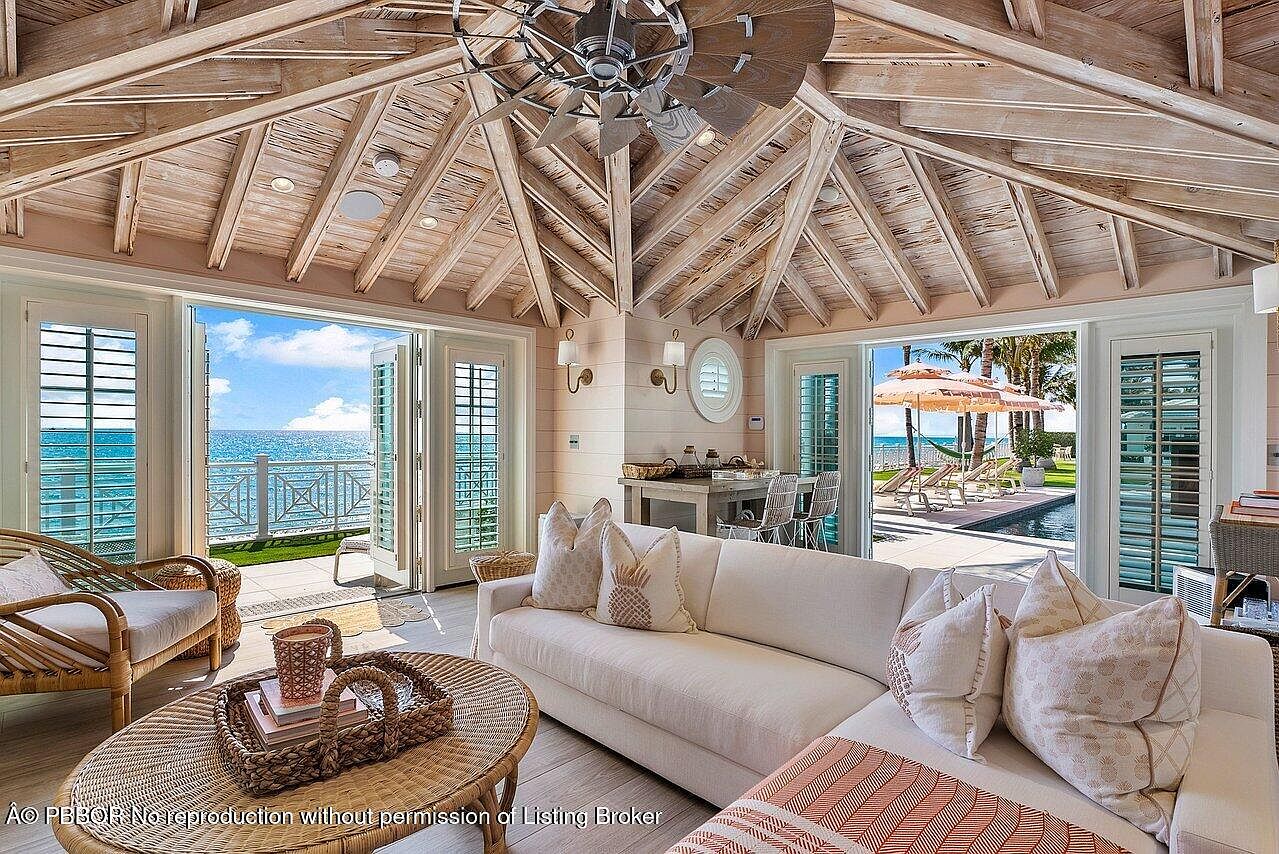 This is an inviting living room with a coastal aesthetic, featuring a vaulted wood ceiling and large bi-fold doors that open to an ocean view. The room is furnished with a comfortable white sofa, wicker chairs, and a round wicker coffee table, creating a relaxed and airy atmosphere. The open design seamlessly blends indoor and outdoor living, perfect for enjoying the seaside setting.