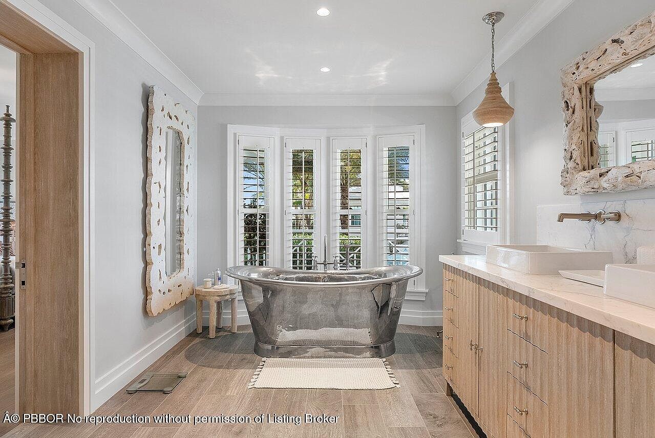 This is a luxurious primary bathroom featuring a freestanding silver bathtub as a focal point, positioned in front of a bay window with plantation shutters. The room includes a double vanity with light wood cabinetry and marble countertops, complemented by unique driftwood-style mirrors. The overall aesthetic is bright, airy, and upscale, creating a spa-like atmosphere.