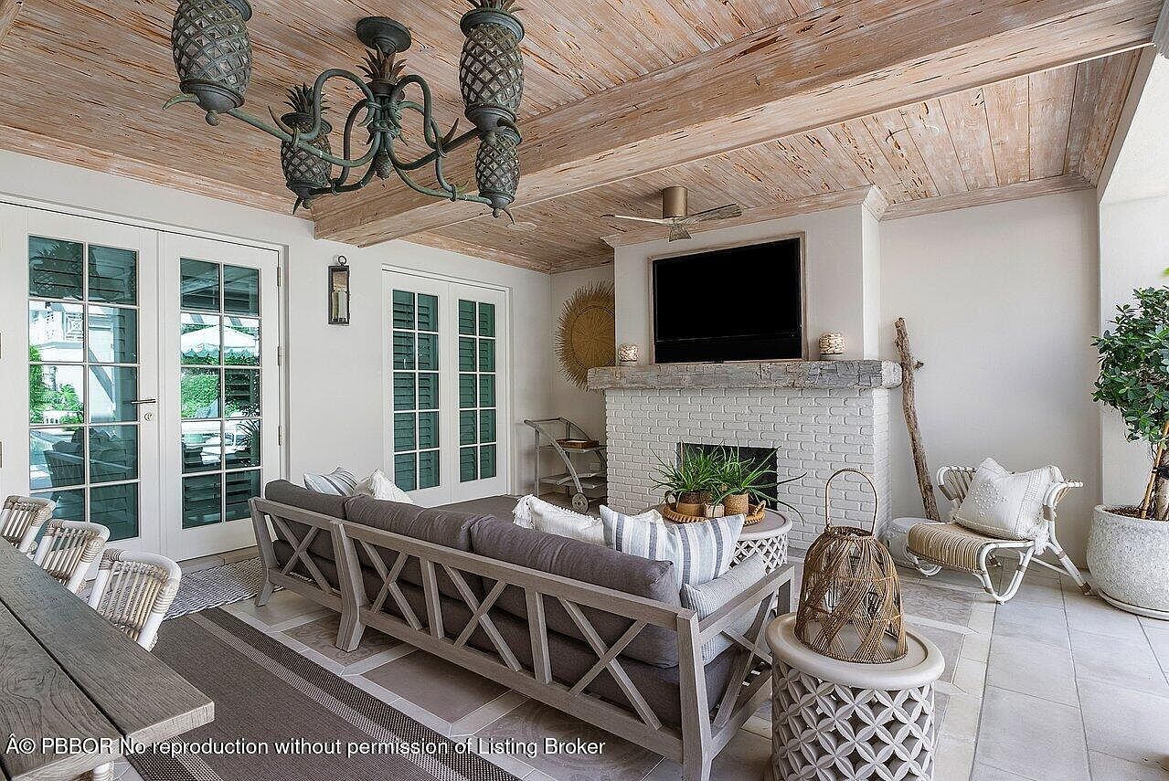 This is a well-decorated outdoor living space featuring a comfortable gray sofa with striped pillows, a white-painted brick fireplace with a mounted TV, and a unique pineapple chandelier. The ceiling is finished with light-colored wood planks, and the space is accessible through glass doors, creating a seamless transition between indoor and outdoor living. The overall impression is one of relaxed elegance and comfort.
