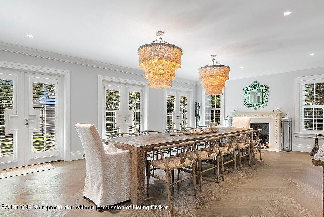 This is an interior shot of a dining room featuring a long wooden table with seating for ten, illuminated by two tiered chandeliers. The room has multiple sets of French doors with shuttered windows, a decorative fireplace with a mirror above, and herringbone wood flooring. The overall impression is elegant and well-lit, suitable for formal dining and entertaining.