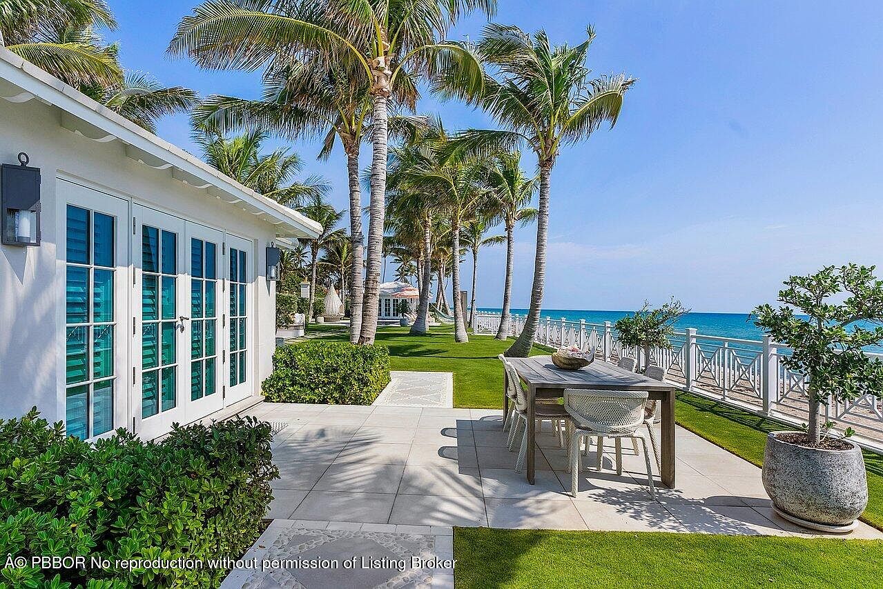 This exterior shot showcases a luxurious patio/deck area with a dining table set for outdoor entertaining. The space features a tiled patio, manicured lawn, and lush landscaping, with palm trees providing shade and a tropical ambiance. A white fence borders the property, offering views of the ocean and beach, creating a serene and upscale outdoor living space.