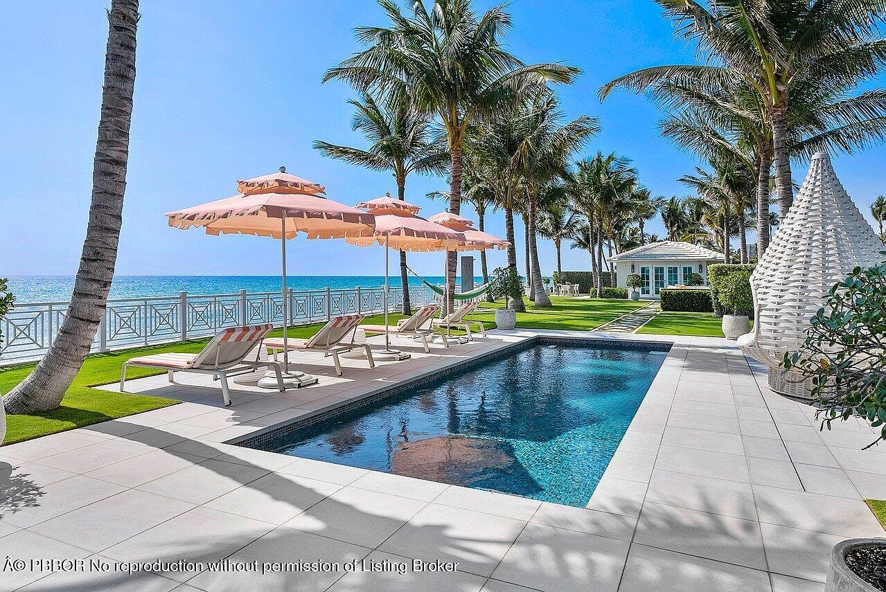 This image showcases a luxurious outdoor pool area with a stunning ocean view. The pool is surrounded by a tiled patio, lounge chairs with umbrellas, and lush palm trees. A white gazebo and manicured lawn add to the property's appeal, creating a serene and upscale atmosphere.