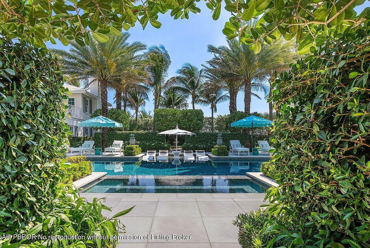 This image showcases a luxurious pool area framed by lush greenery and palm trees. The pool features a dark blue tile interior, complemented by light-colored lounge chairs and umbrellas. The overall impression is one of tranquility and upscale outdoor living, perfect for relaxation and entertainment.