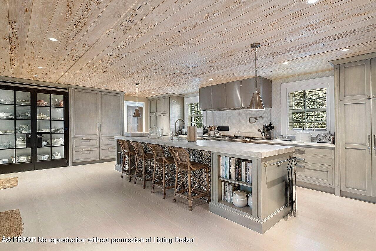 This is a well-lit kitchen featuring a large island with seating, custom cabinetry, and stainless steel appliances. The island has a unique patterned backsplash and built-in shelving. The ceiling is wood paneled, adding a rustic touch to the otherwise modern design.