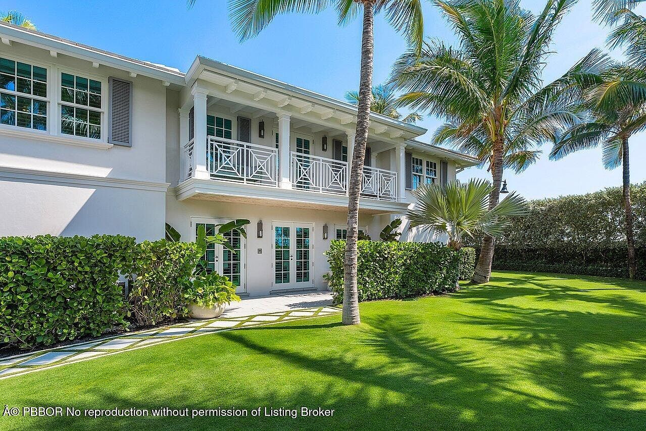 This is a front exterior view of a two-story white house with a balcony. The house is surrounded by lush green lawn and manicured hedges, with palm trees adding a tropical feel. The architecture features clean lines and a symmetrical design, suggesting a sophisticated and well-maintained property.