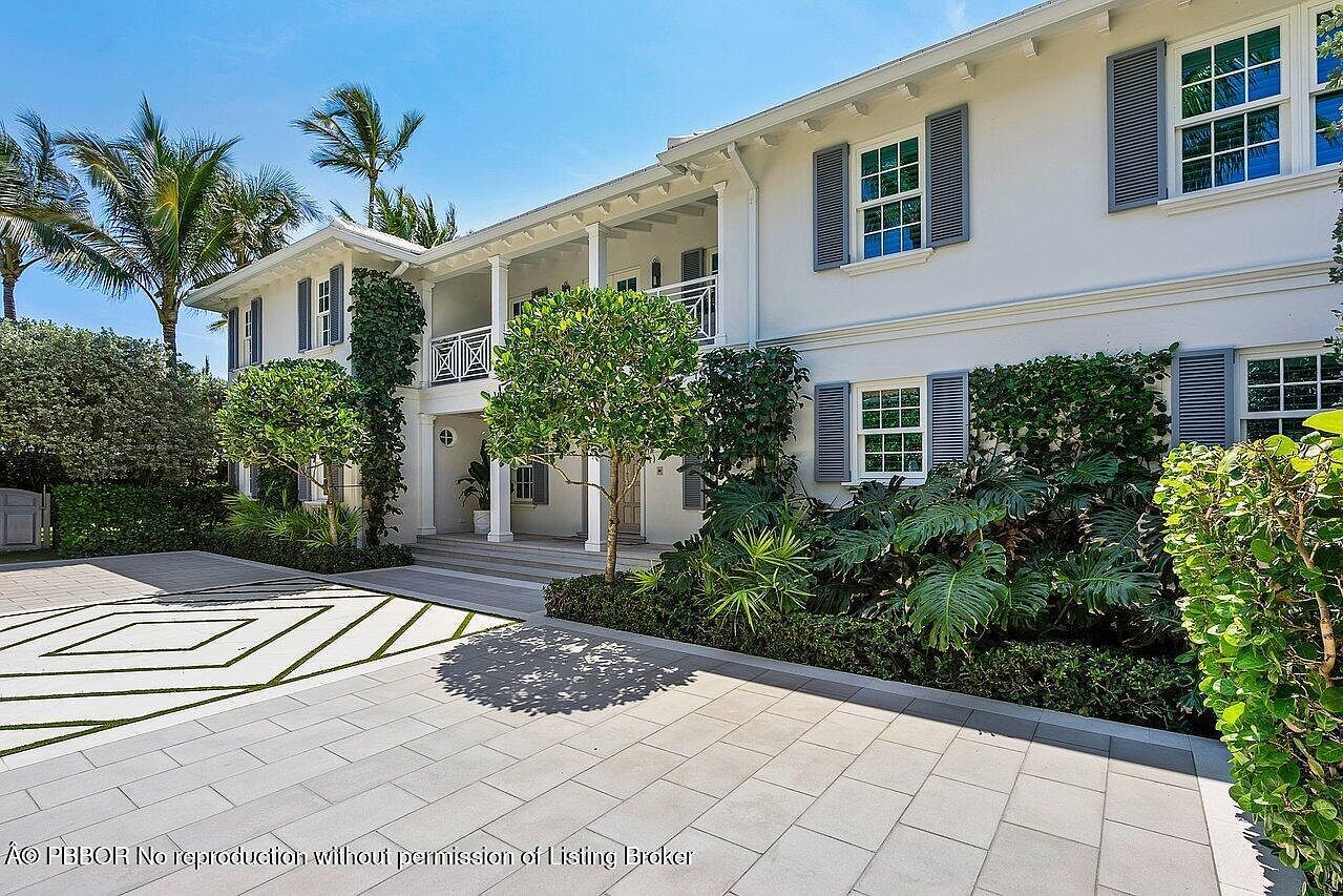 This is a front exterior view of a two-story white house with gray shutters and a second-story balcony. The house is surrounded by lush greenery, including palm trees and manicured shrubs. The driveway is paved with light-colored stones arranged in a geometric pattern, creating a welcoming and luxurious entrance.