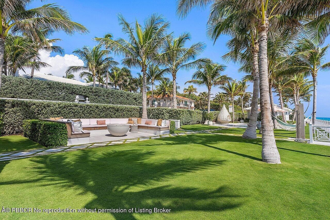 This image showcases a beautifully landscaped yard with lush green grass, mature palm trees, and manicured hedges. A built-in seating area with a fire pit provides an inviting outdoor space for relaxation and entertainment. The overall impression is one of luxury and tranquility, perfect for enjoying the tropical surroundings.