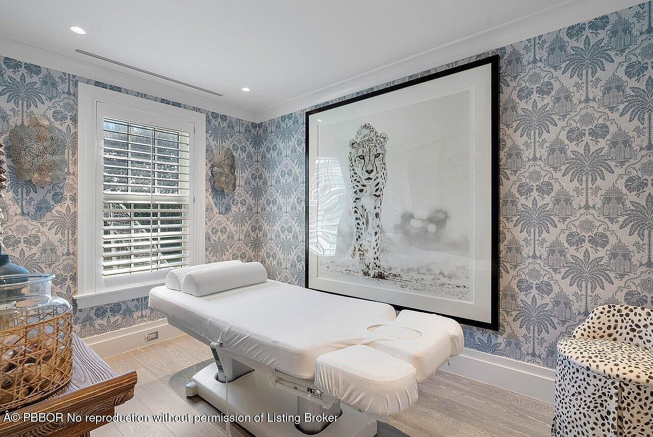 This interior shot showcases a serene room featuring a massage table as its focal point, suggesting a spa-like atmosphere. The walls are adorned with patterned wallpaper in muted blue tones, complemented by a large framed print of a cheetah. Natural light filters through the window, enhancing the room's tranquil ambiance.