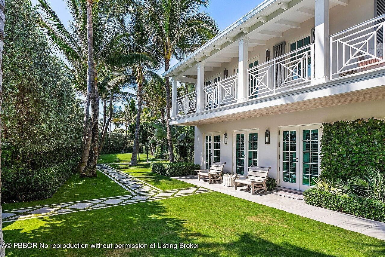 This image showcases a beautifully landscaped yard with a luxurious two-story home in the background. The yard features lush green grass, palm trees, and a decorative stone pathway. The home has a white exterior with a balcony and multiple French doors, creating an inviting and elegant atmosphere.
