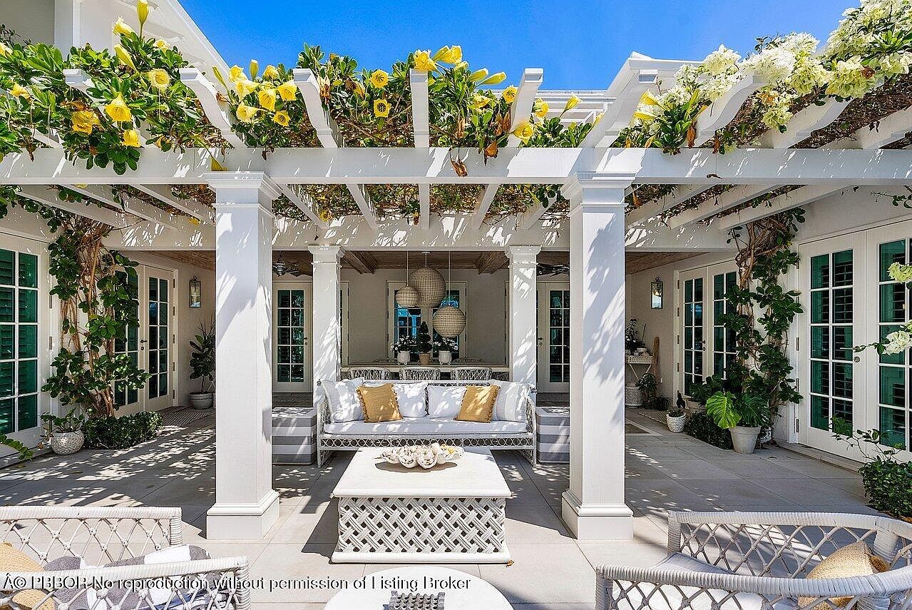 This is a beautifully designed outdoor patio area featuring a white pergola adorned with flowering vines, providing shade and a touch of elegance. The space is furnished with comfortable wicker seating, including a sofa with decorative pillows and chairs, arranged around a stylish coffee table. The patio is paved with light-colored stone, creating a serene and inviting atmosphere perfect for relaxation and entertaining.