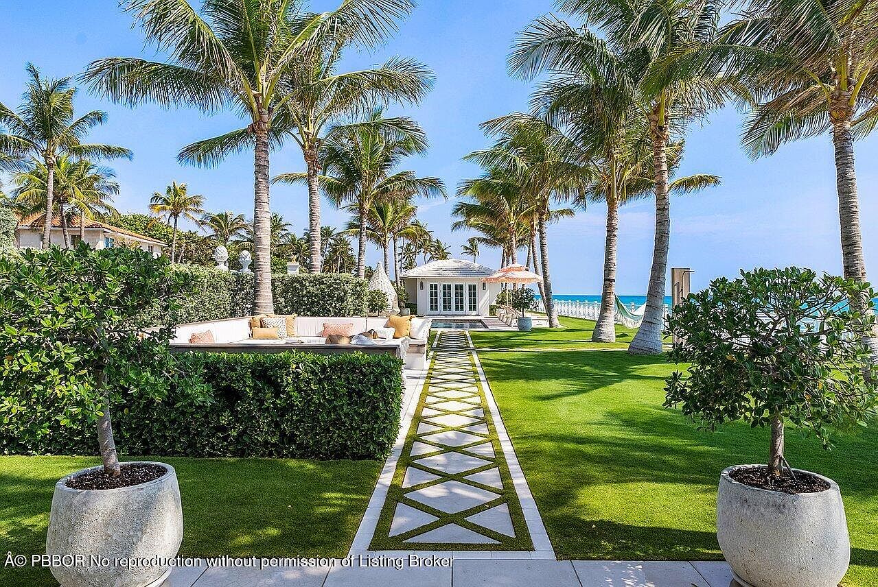 This image showcases a meticulously landscaped yard featuring a geometric stone pathway leading to a charming white cabana. Lush green lawns are bordered by mature palm trees and manicured hedges, creating a serene and luxurious outdoor space. The scene evokes a sense of tranquility and high-end living.