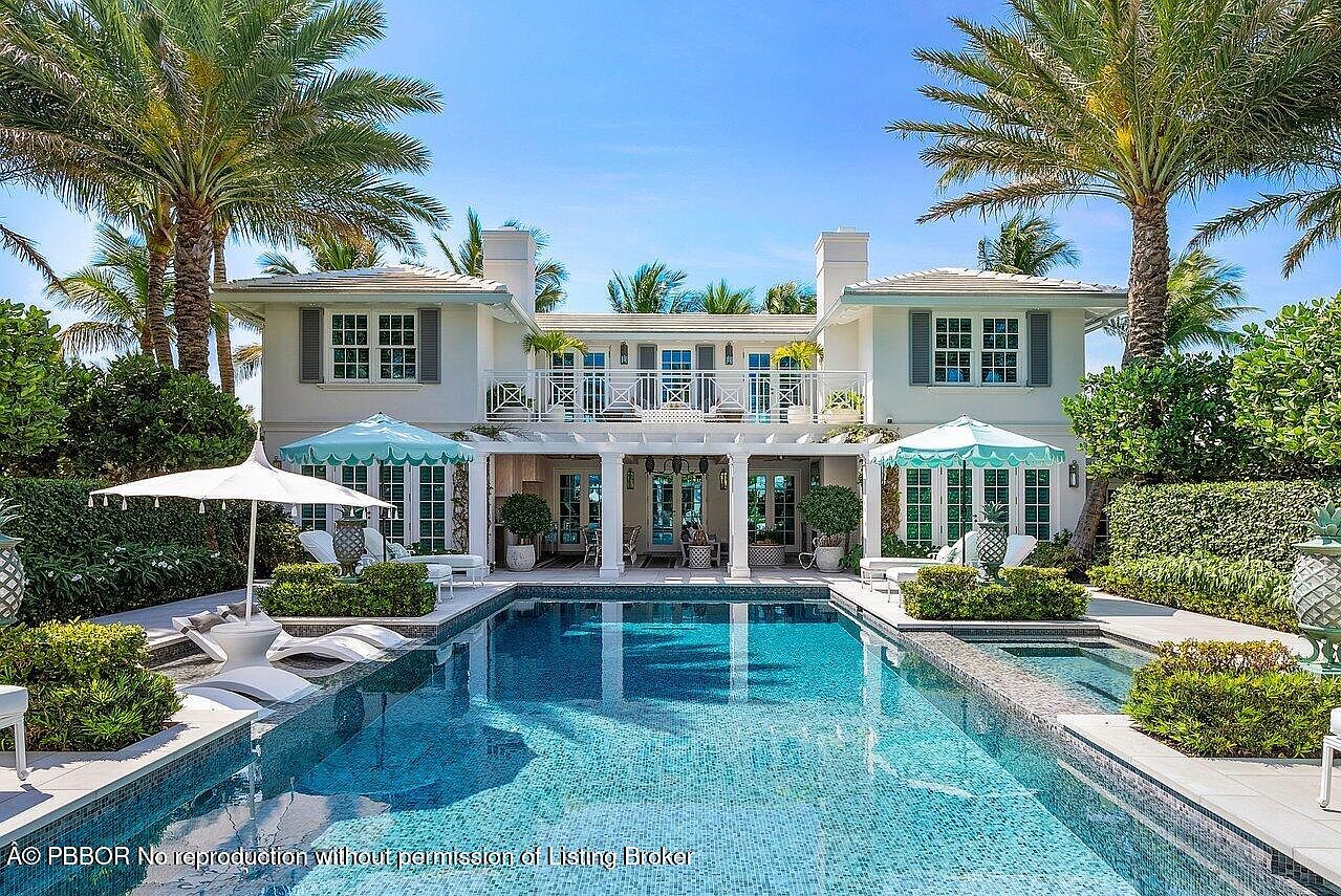 This image showcases a luxurious backyard featuring a large, rectangular swimming pool with a mosaic tile finish. The pool is surrounded by manicured greenery, lounge chairs, and umbrellas, creating a relaxing oasis. The backdrop is a grand, white two-story house with a balcony, adding to the overall elegance and appeal of the property.