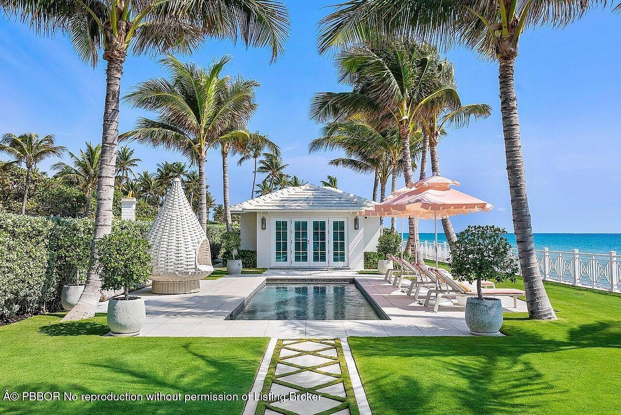 This image showcases a luxurious outdoor pool area, framed by lush palm trees and manicured green lawns. A charming pool house with French doors sits at the pool's edge, complemented by stylish lounge chairs and a decorative umbrella. The scene evokes a sense of tranquility and upscale coastal living.