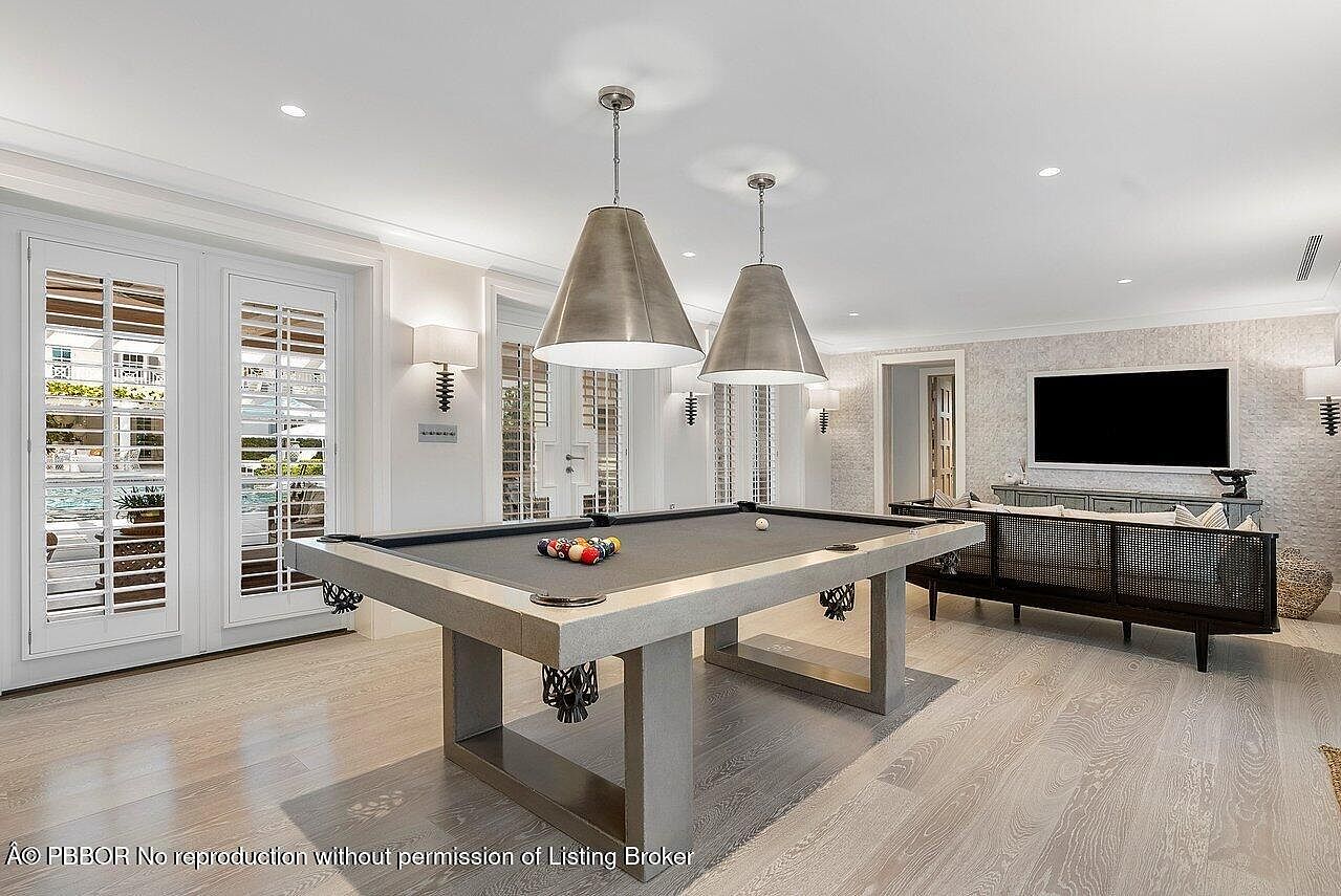 This interior shot showcases a well-lit living room featuring a modern pool table as a central element. The room is decorated in neutral tones with light wood flooring, complemented by two large pendant lights hanging above the pool table. A console table with a television and decorative items is visible in the background, adding to the room's sophisticated and recreational atmosphere.