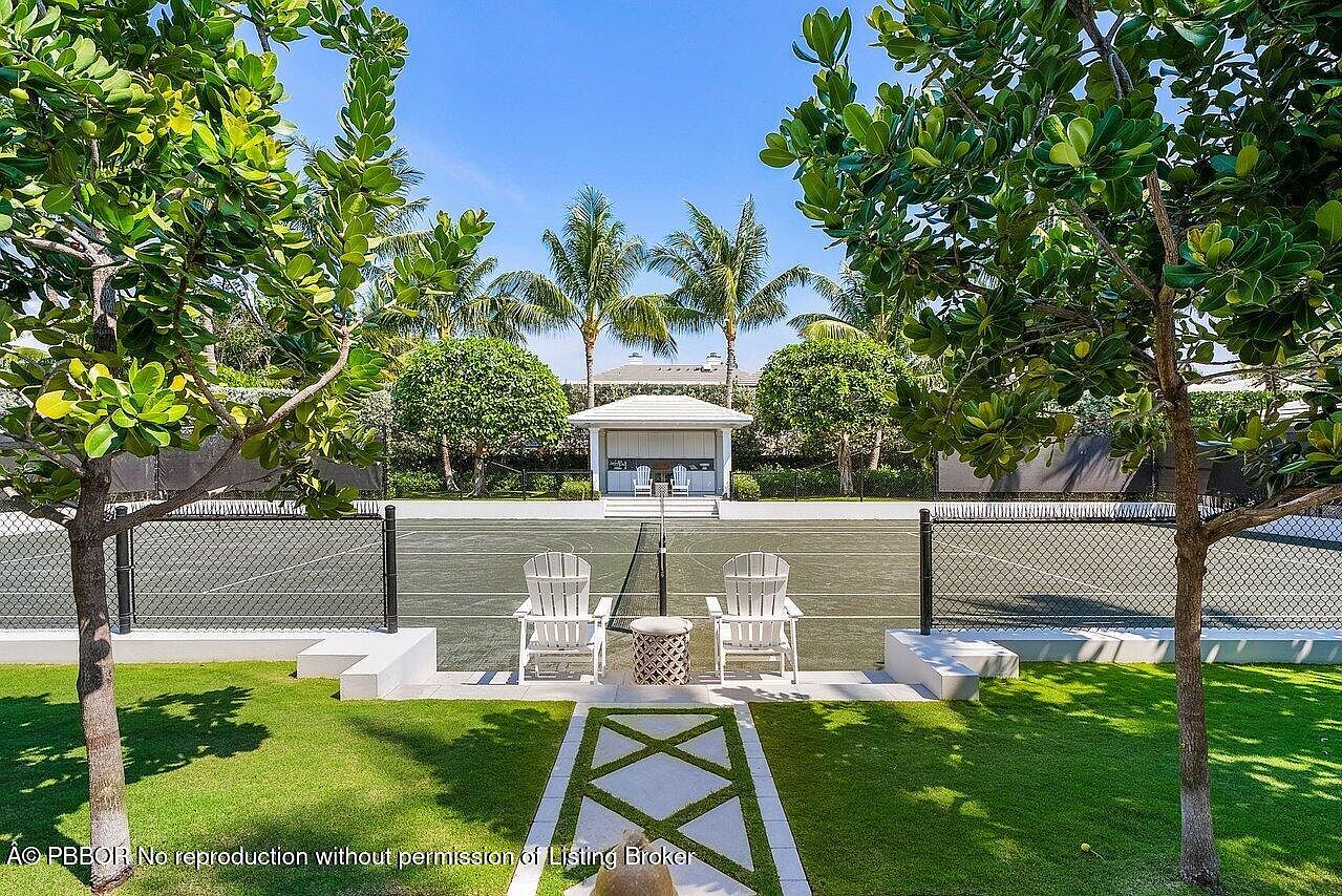 This image showcases a beautifully landscaped yard featuring a tennis court as its focal point. Two white Adirondack chairs are positioned near the court, creating an inviting space for relaxation. The lush green lawn, framed by trees and a decorative walkway, enhances the property's appeal and offers a serene outdoor setting.
