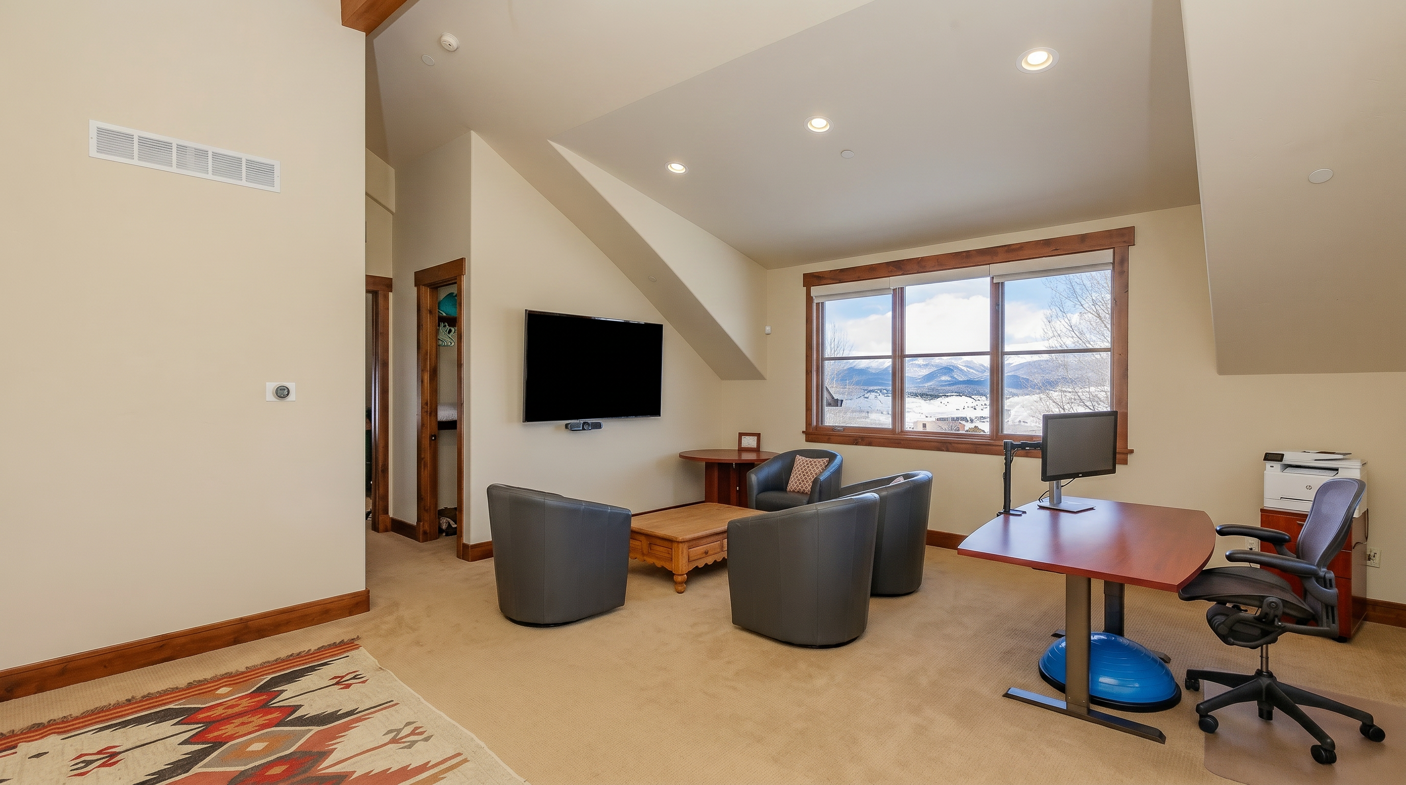 This versatile room functions as a combined home office and lounge area, featuring a spacious desk setup, a comfortable seating arrangement with three leather armchairs, and a wall-mounted television. The space is characterized by its vaulted ceilings, neutral carpeting, and a large window that provides a stunning, panoramic view of snow-capped mountains. The overall atmosphere is professional yet relaxed, making it an ideal space for both productivity and leisure.