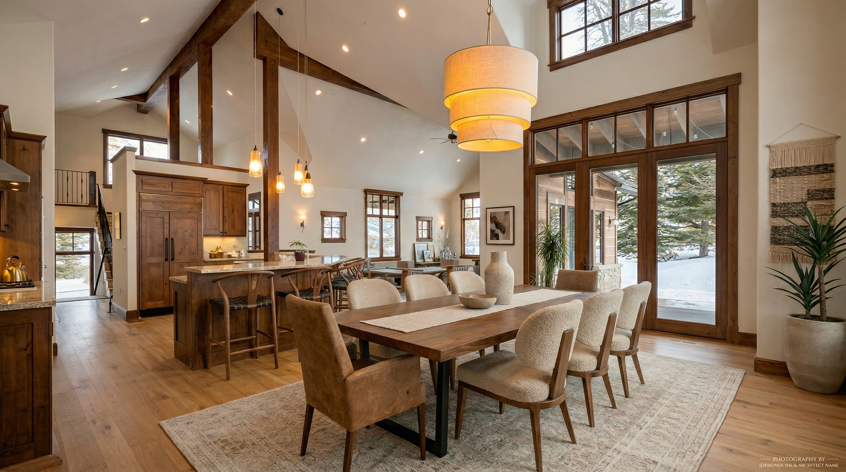 This spacious, open-concept dining area features a large wooden table with upholstered chairs, set beneath a striking tiered pendant light fixture. The room flows seamlessly into a well-appointed kitchen with a breakfast bar, all under a vaulted ceiling with exposed wooden beams. Large windows and glass doors provide abundant natural light and a view of the snowy exterior, creating a warm and inviting atmosphere.