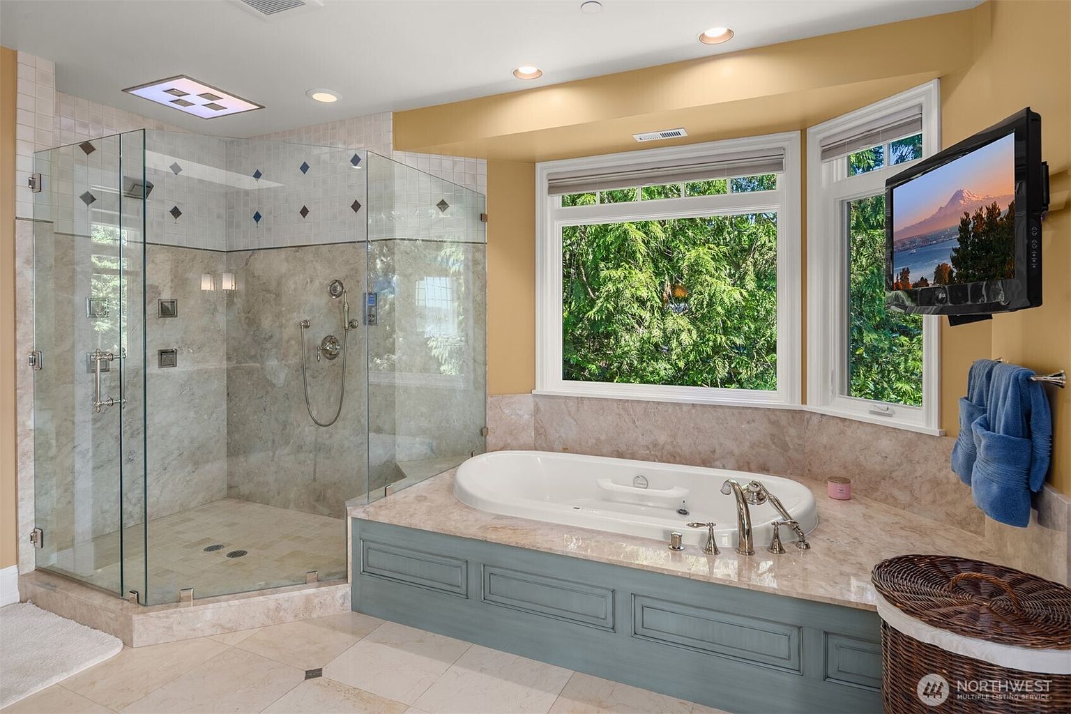 This luxurious primary bathroom features a spacious glass-enclosed walk-in shower with intricate tile work and a deep soaking tub set into a marble-topped vanity. A large bay window provides ample natural light and a scenic view, while a wall-mounted television adds a modern touch of convenience. The room is finished with elegant cabinetry and neutral tones, creating a serene and spa-like atmosphere.