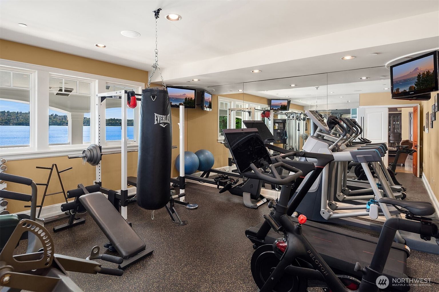 This well-equipped home gym features a variety of fitness equipment, including a Peloton bike, a treadmill, a weight bench, and a heavy punching bag. The room is lined with a large wall mirror that enhances the sense of space, while expansive windows provide a stunning, unobstructed view of the lake. The overall atmosphere is professional and motivating, perfect for a high-end residential fitness area.