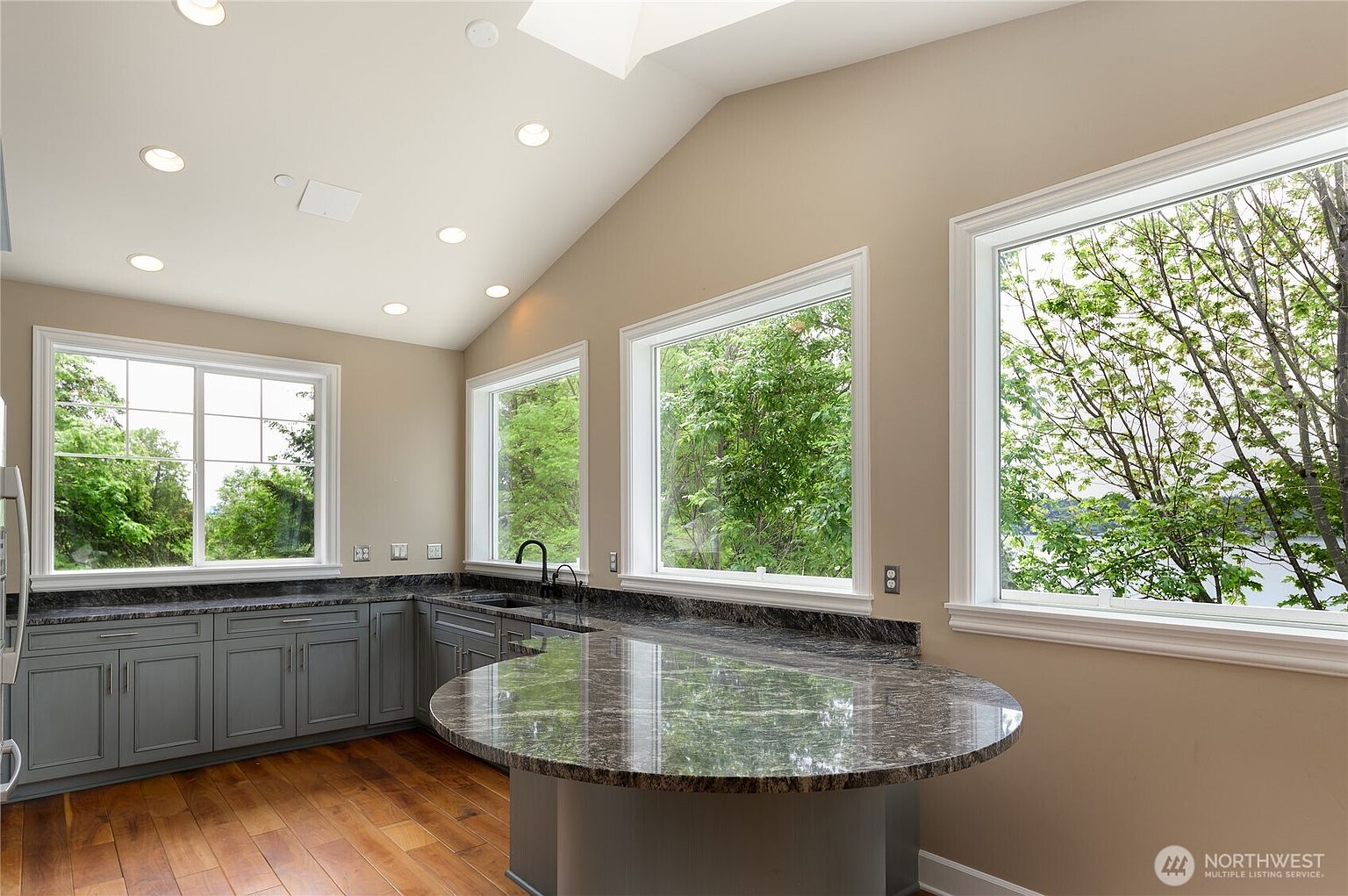 This bright and airy kitchen features sleek grey cabinetry paired with dark, polished granite countertops and a unique curved peninsula island. Large windows wrap around the space, offering expansive views of the lush greenery and water outside, while recessed lighting illuminates the vaulted ceiling. The warm hardwood flooring provides a beautiful contrast to the cool tones of the cabinetry, creating a sophisticated and inviting atmosphere.