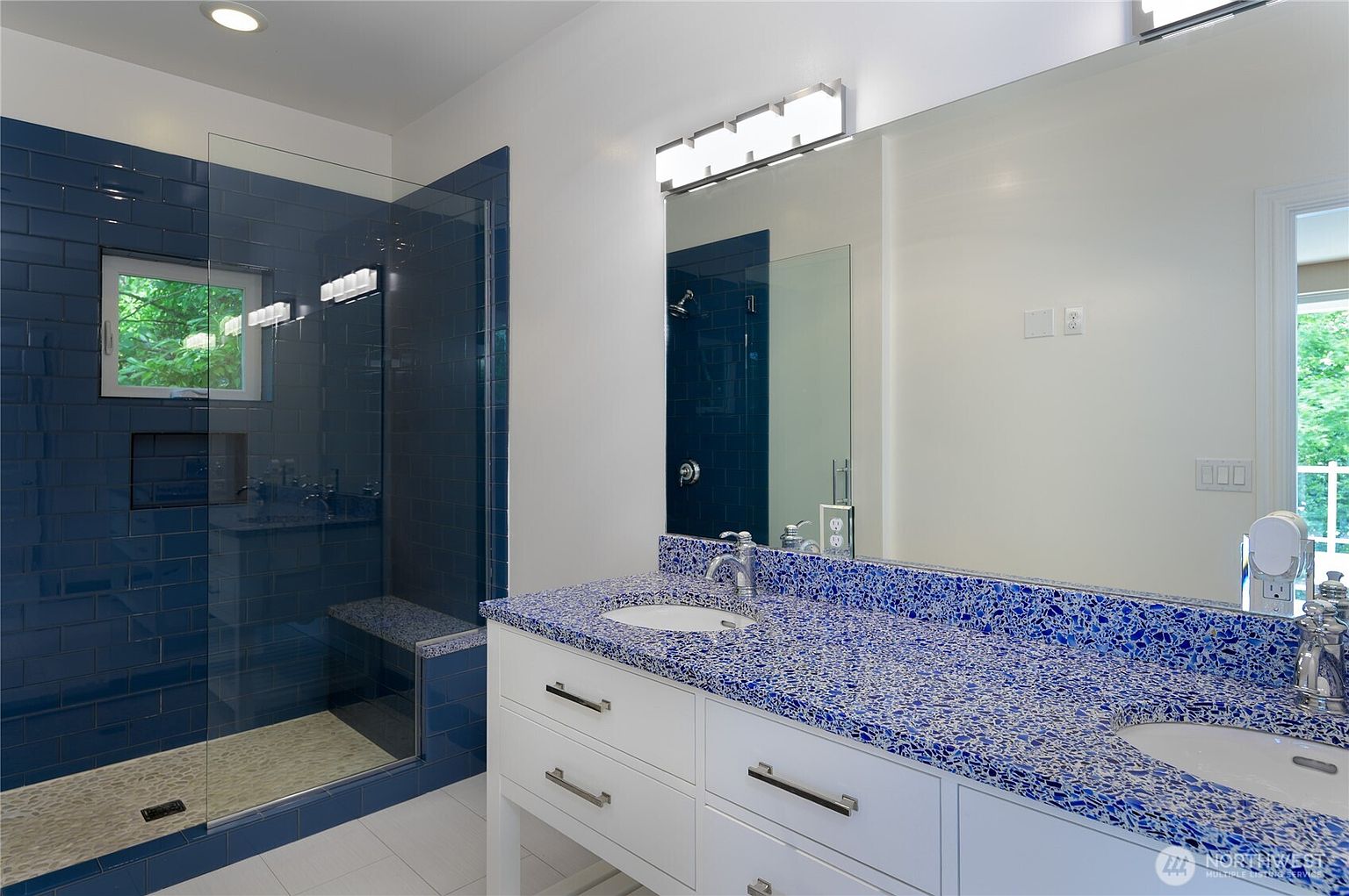 This modern primary bathroom features a striking blue-and-white speckled countertop paired with a crisp white double vanity. A spacious walk-in shower with deep blue subway tiles and a glass enclosure creates a bold, contemporary aesthetic. The large mirror and bright lighting enhance the sense of space, while the clean lines and vibrant color palette offer a refreshing and stylish atmosphere.