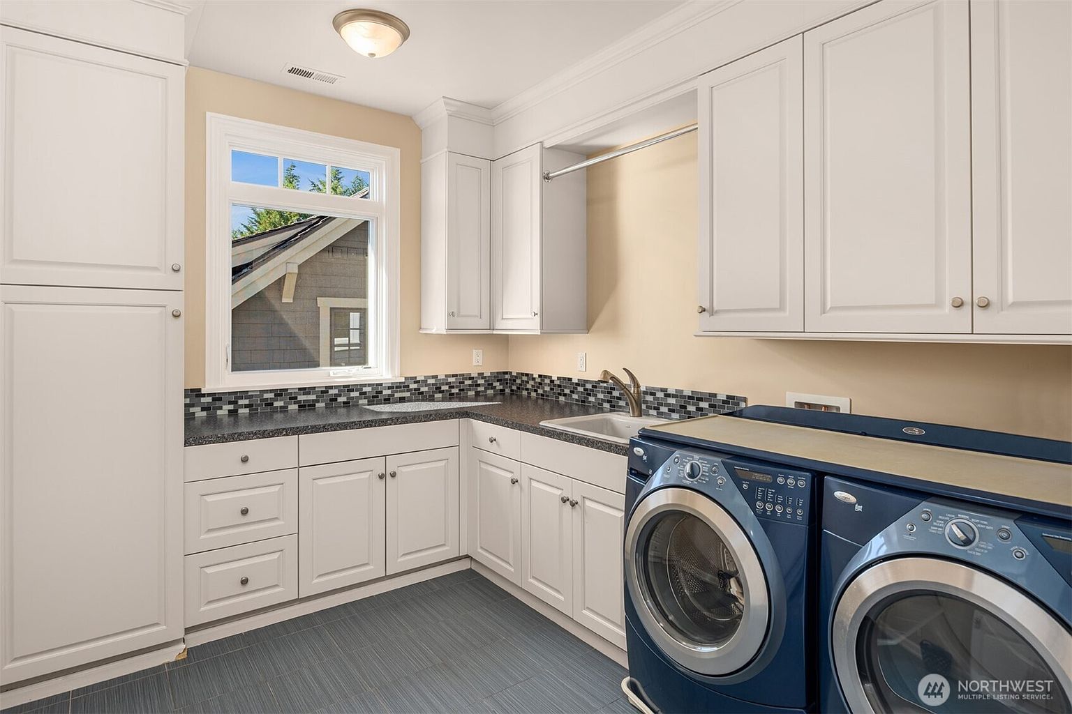 This bright and functional laundry room features crisp white cabinetry, dark countertops, and a mosaic tile backsplash. The space is equipped with a deep utility sink and modern blue front-loading washer and dryer units, all set against a sleek, dark-toned tiled floor. A large window allows natural light to fill the room, creating an inviting and organized workspace.