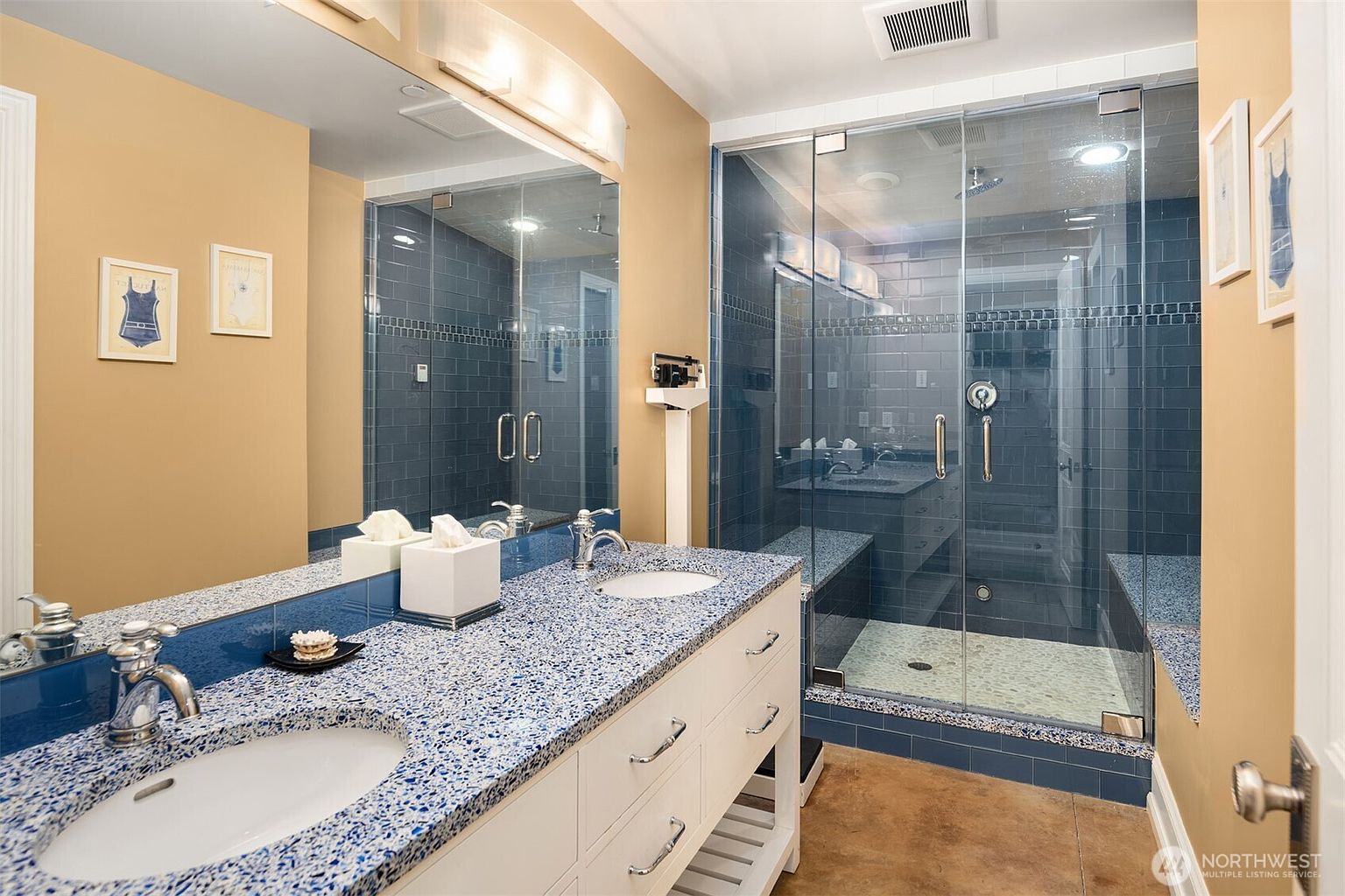 This primary bathroom features a dual-sink vanity with a distinctive blue-flecked granite countertop and white cabinetry. A spacious walk-in shower with blue subway tiles, a pebble-stone floor, and a built-in bench serves as the focal point. The room is accented with warm-toned walls and framed artwork, creating a clean and functional aesthetic.