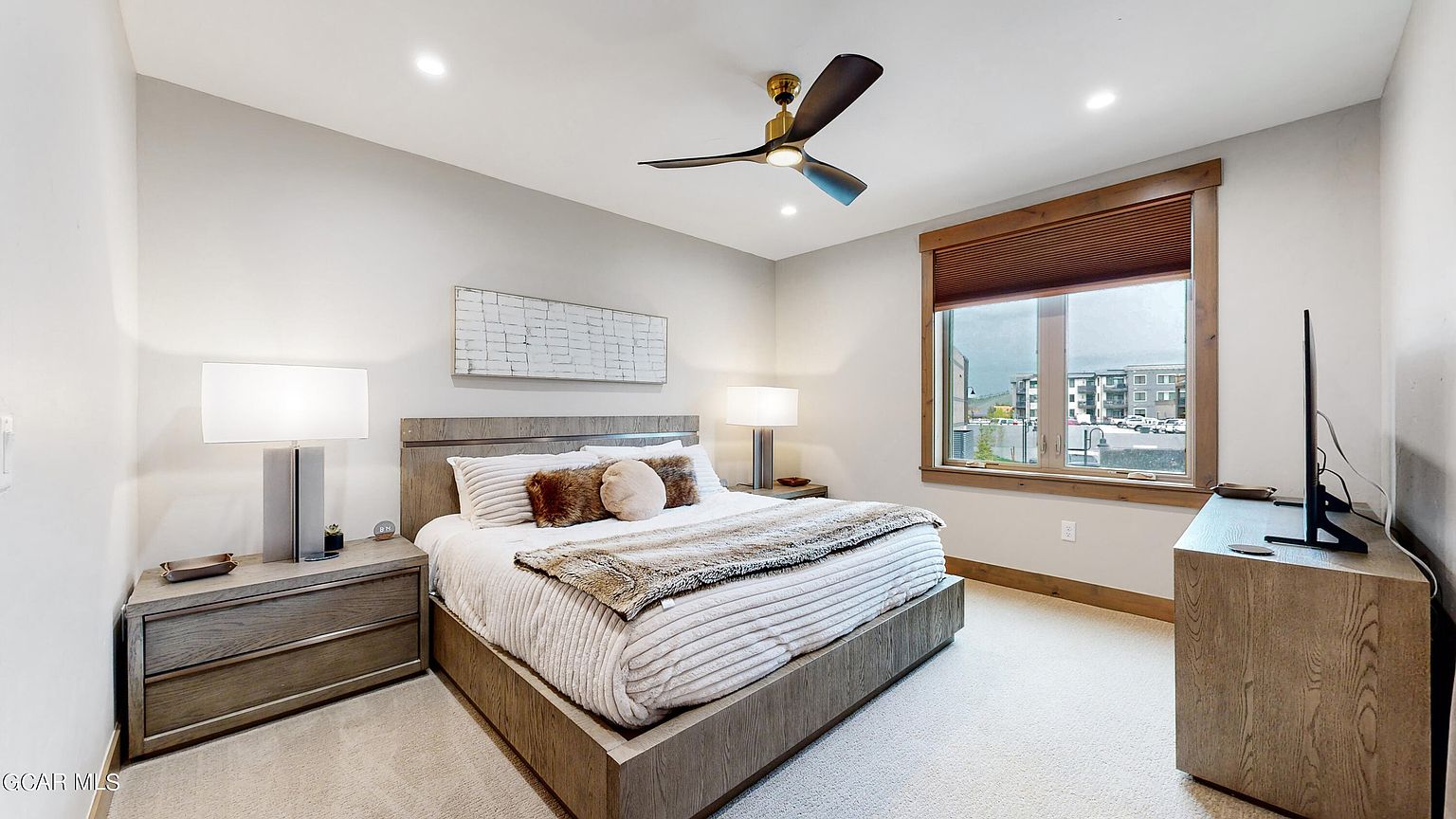 This is a well-lit primary bedroom featuring a king-size bed with a light wood frame, adorned with plush bedding and decorative pillows. The room includes matching nightstands with lamps, a ceiling fan, and a window offering natural light and a view of the outside. A wooden dresser with a TV completes the space, creating a comfortable and inviting atmosphere.