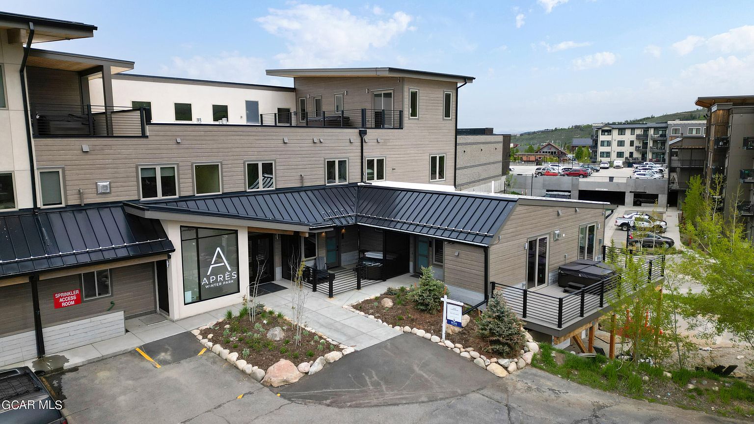This is a front exterior view of a modern multi-story building with a combination of light brown siding and black metal roofing. The building features a well-maintained landscape with rocks and small trees, a paved walkway leading to the entrance, and a sign with the building's name. The overall impression is clean, contemporary, and inviting.