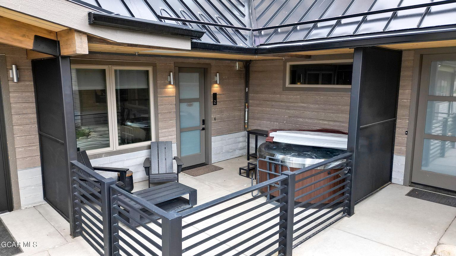 This image showcases a modern patio area featuring a hot tub, sleek black metal railings, and contemporary outdoor furniture. The patio is partially covered by a metal roof, providing shade and protection. The exterior of the building is clad in horizontal wood siding, adding to the modern aesthetic.