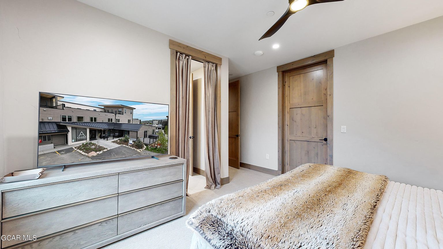 This is a primary bedroom featuring a light wood dresser with a television on top displaying an exterior shot of a building. The room has neutral-toned walls, a ceiling fan, and a bed with a textured blanket. There are two wooden doors, one with a curtain, adding to the room's character.
