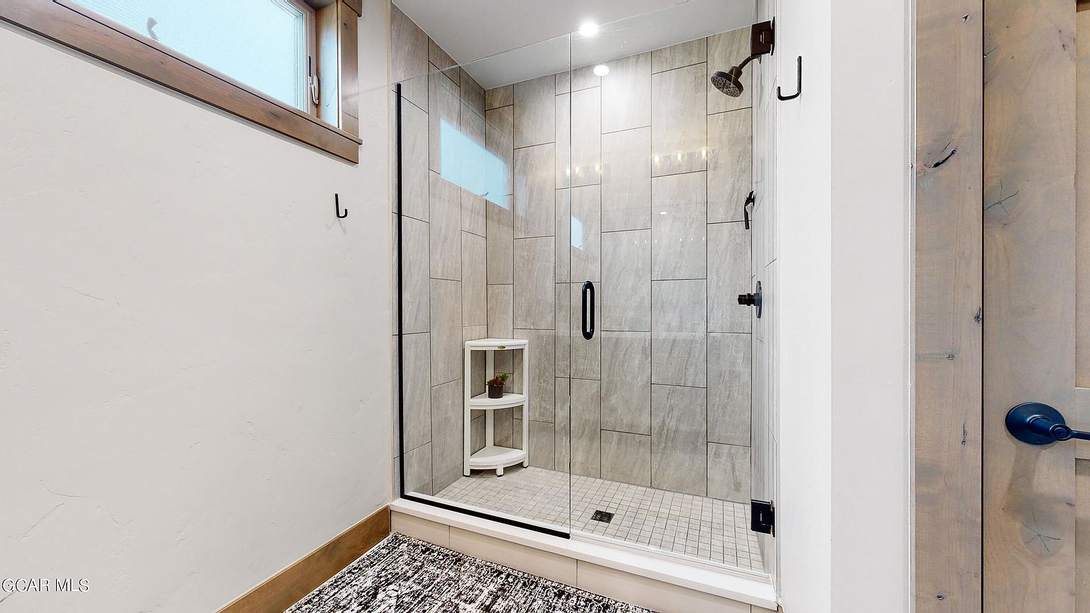 This is a well-lit bathroom featuring a modern glass-enclosed shower with gray tiled walls and a tiled floor. A small white corner shelf sits inside the shower, and black hardware accents the glass door and shower fixtures. The overall impression is clean and contemporary.
