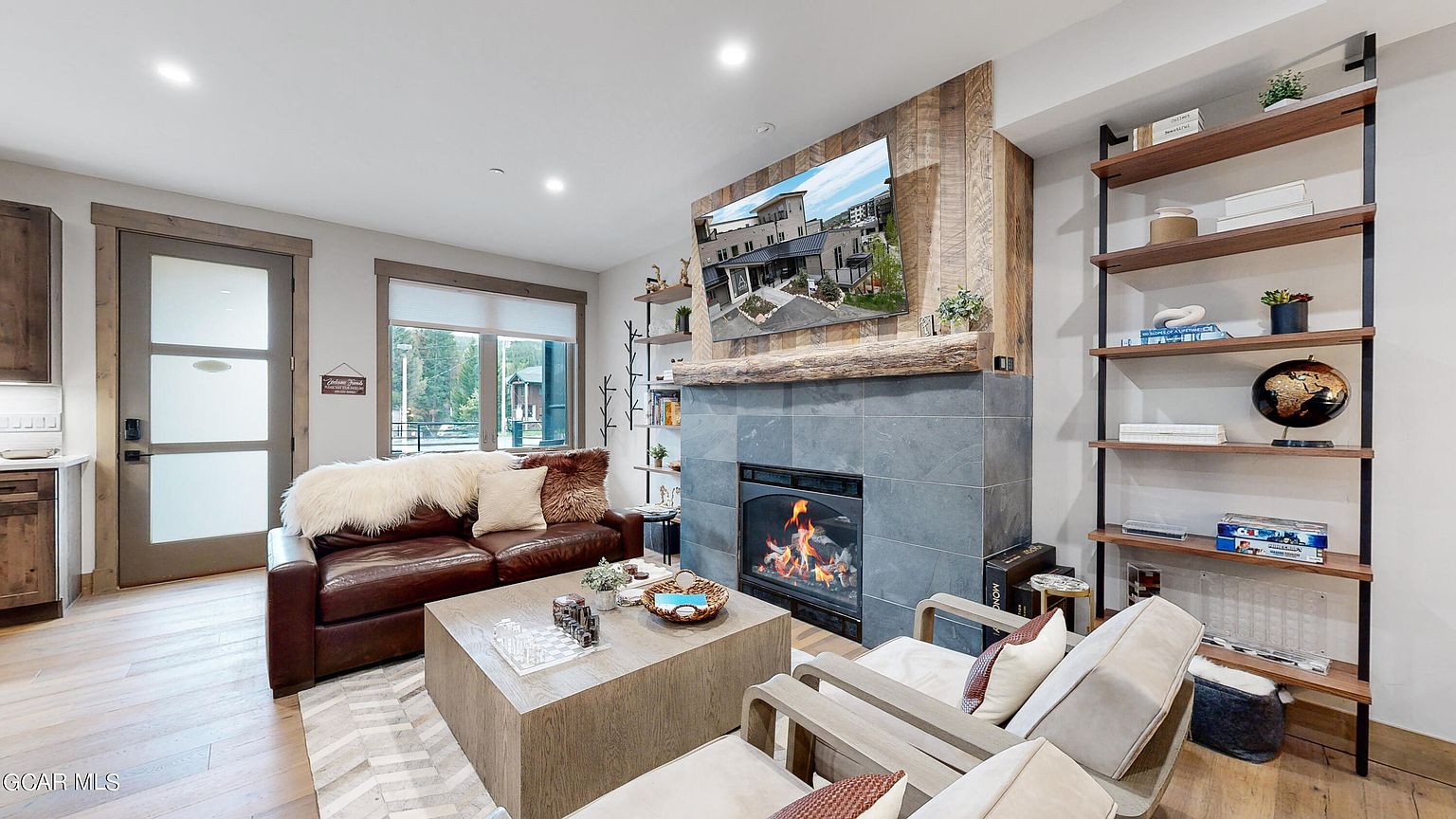This is an interior shot of a living room featuring a stone fireplace with a wooden mantel and a mounted television. The room is furnished with a leather sofa, two armchairs, and a large square coffee table. Natural light streams in through a window, and the overall style is rustic and cozy.