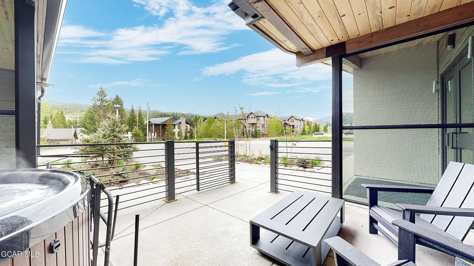 This image showcases a modern patio or balcony area, featuring sleek horizontal railing, contemporary outdoor furniture including a table and chairs, and a hot tub. The space offers an inviting outdoor living area with views of the surrounding neighborhood and distant mountains, blending indoor and outdoor living seamlessly.