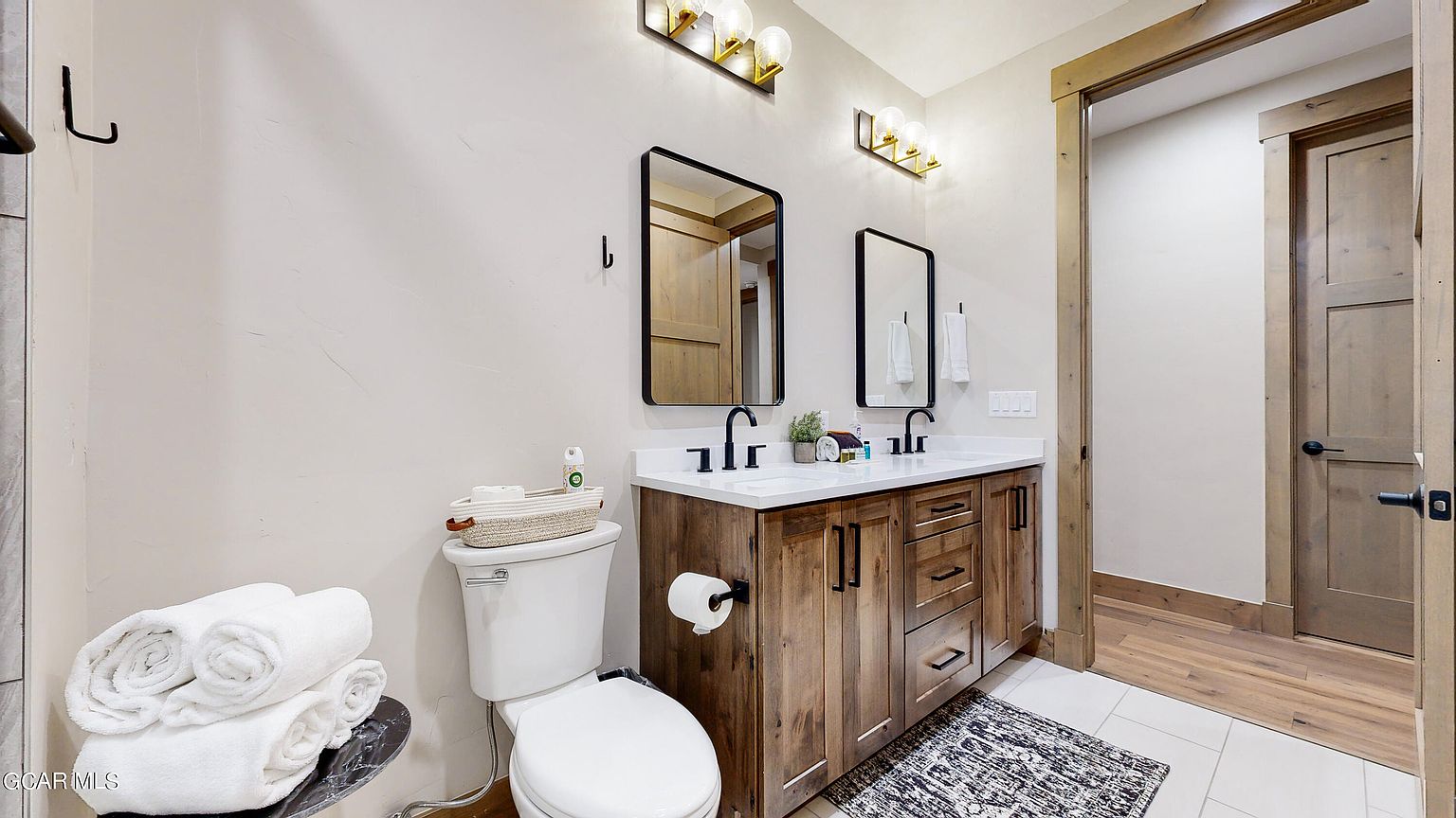This is a well-lit bathroom featuring a double vanity with a white countertop and rustic wood cabinetry. Two black-framed mirrors hang above the sinks, complemented by modern light fixtures. A toilet sits to the left, and a doorway with a wooden frame and door is visible on the right, adding to the rustic-modern aesthetic.