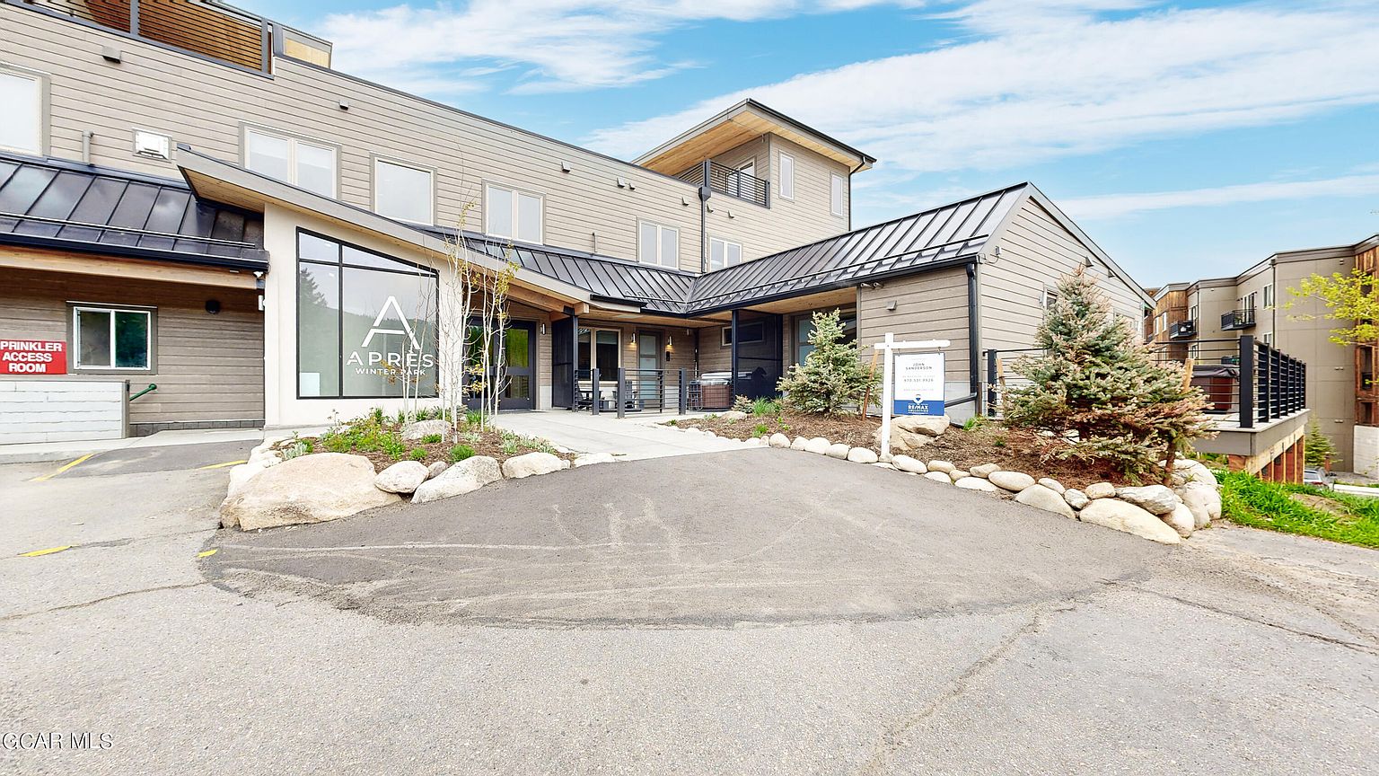 This is a front view of a modern multi-story building with a combination of light brown siding and black metal roofing. The building features large windows, a well-maintained landscape with rocks and small trees, and a paved driveway leading up to the entrance. A real estate sign is visible, indicating the property is for sale.