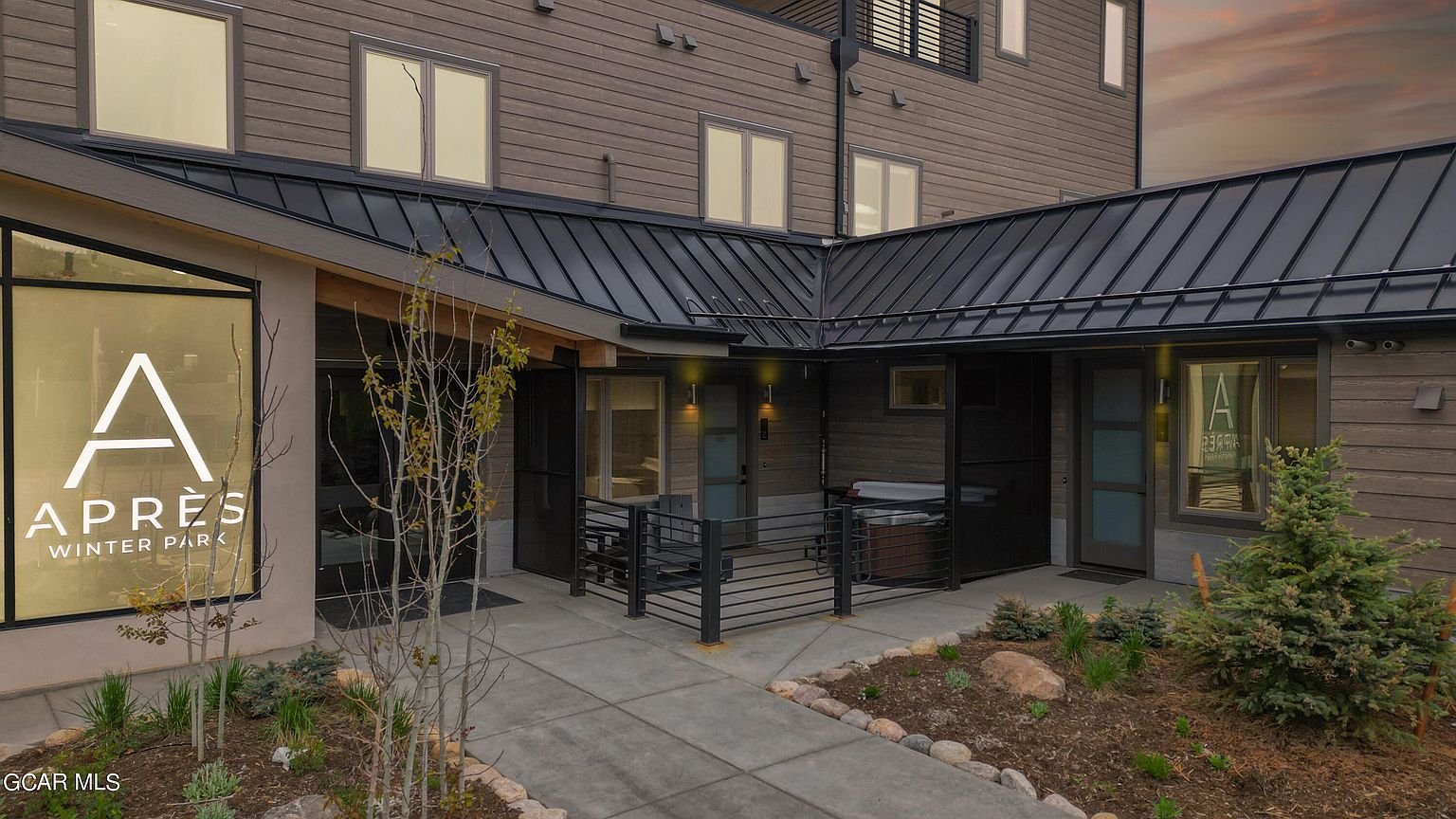 The image showcases the entryway of a modern building, possibly a condominium or townhome complex. The exterior features a combination of wood siding and a dark metal roof, creating a contemporary aesthetic. A well-maintained landscape with small trees and stone accents adds to the curb appeal, while the 'Apres Winter Park' signage suggests a location in a winter resort area.