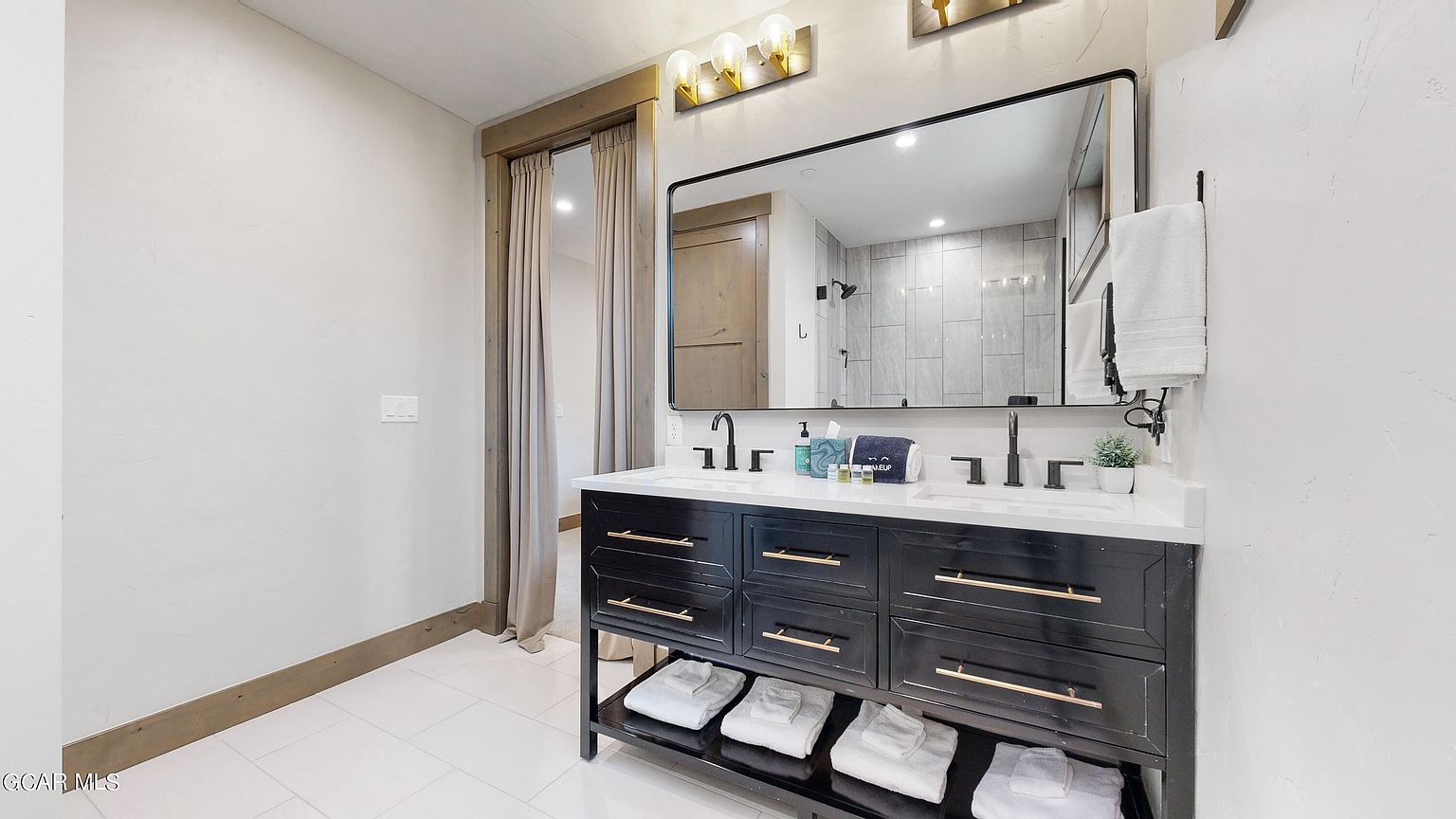 This is a well-lit primary bathroom featuring a double vanity with a black cabinet and gold hardware, topped with a white countertop. A large mirror reflects the shower area, and modern light fixtures illuminate the space. The overall impression is clean, modern, and luxurious.