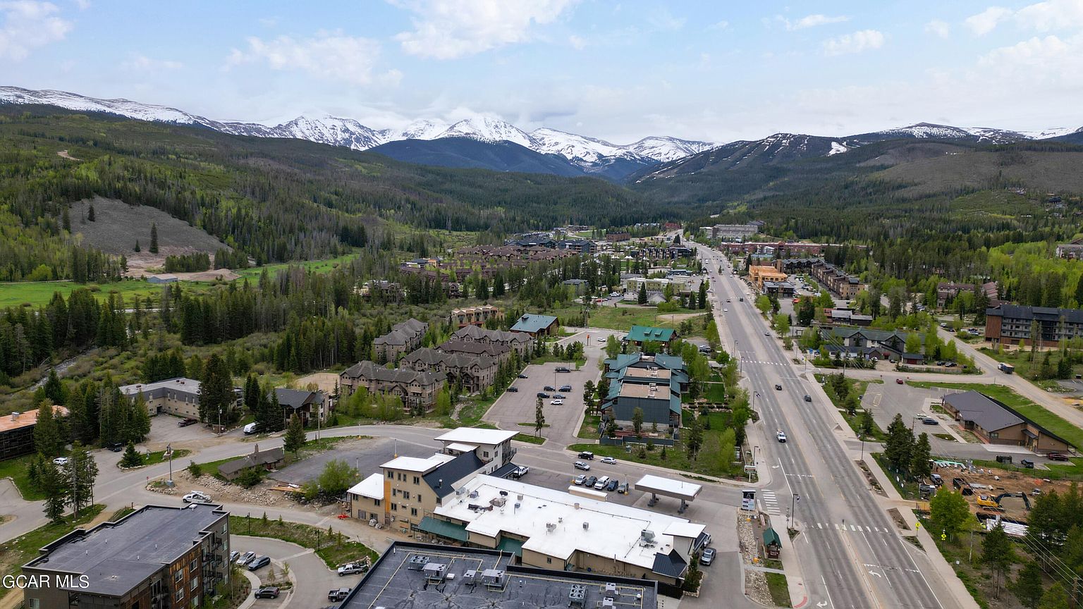 This aerial view showcases a charming town nestled amidst lush green forests and majestic snow-capped mountains. The town features a mix of residential and commercial buildings, with a prominent road leading through the center. The overall impression is one of a peaceful and scenic mountain community, perfect for those seeking a tranquil lifestyle.