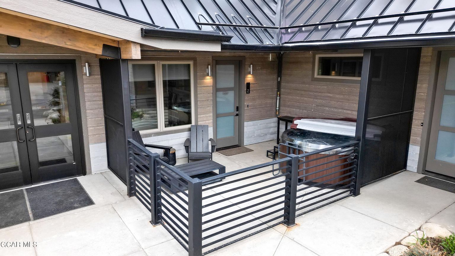 This image showcases a modern patio area featuring a hot tub enclosed by a sleek, horizontal-slat metal railing. The patio includes outdoor seating with a small table, and the area is accessed by a glass-paneled door and a set of double doors, creating an inviting outdoor living space. The exterior is clad in wood-look siding, complemented by a metal roof, adding to the contemporary aesthetic.