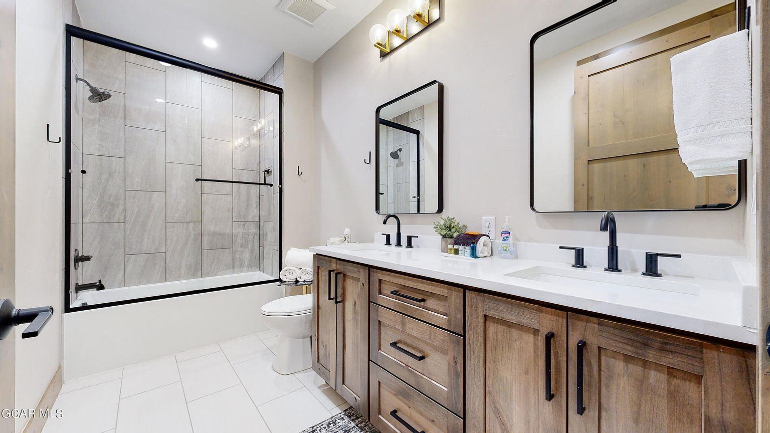 This is a well-lit primary bathroom featuring a double vanity with wooden cabinets and black hardware, topped with a white countertop and dual sinks. Above the vanity are two framed mirrors, and a shower/tub combination with a glass enclosure is visible to the left. The floor is tiled in a light color, contributing to the clean and modern aesthetic of the space.