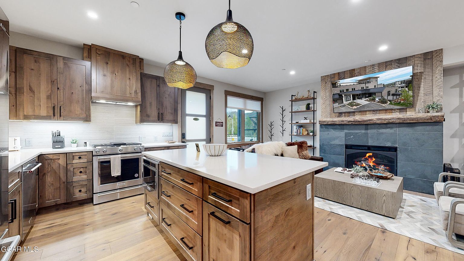 This is an interior shot of a modern kitchen featuring wooden cabinetry and a large island with a white countertop. The kitchen is well-lit by pendant lights and natural light from a window. In the background, there is a living area with a fireplace and a television, creating an open-concept living space.