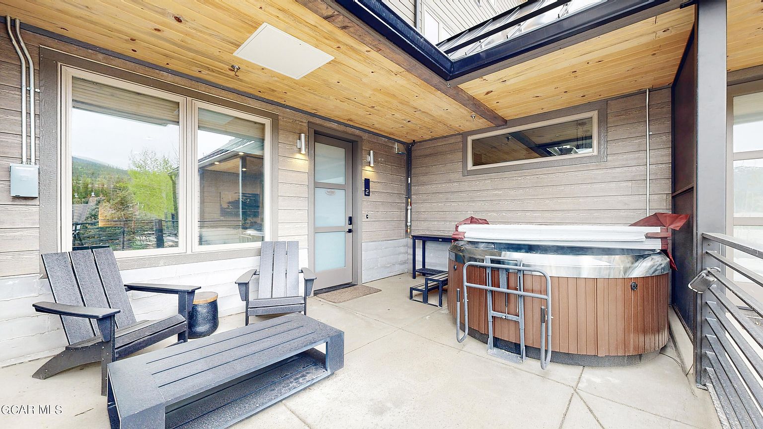 This image showcases a well-appointed outdoor patio or balcony area. It features comfortable seating arrangements, including Adirondack chairs and a bench, alongside a hot tub. The space is covered by a wooden ceiling, providing shelter, and the overall design suggests a relaxing and private outdoor retreat.