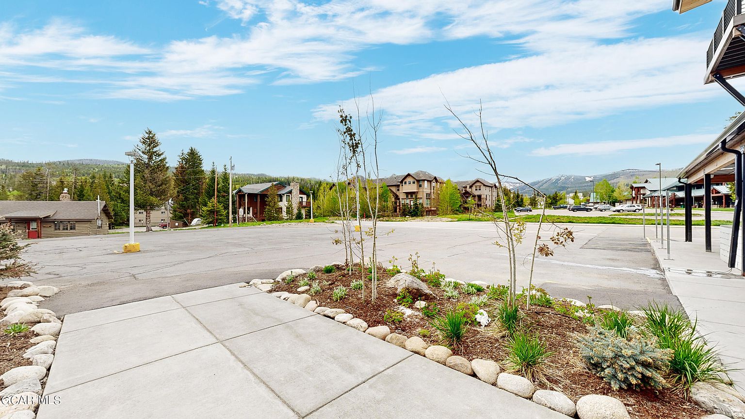 This image showcases the front view of a property, featuring a well-maintained landscape with rocks and plants in the foreground. The building is set against a backdrop of trees and mountains under a partly cloudy sky. The overall impression is one of a peaceful and scenic location.